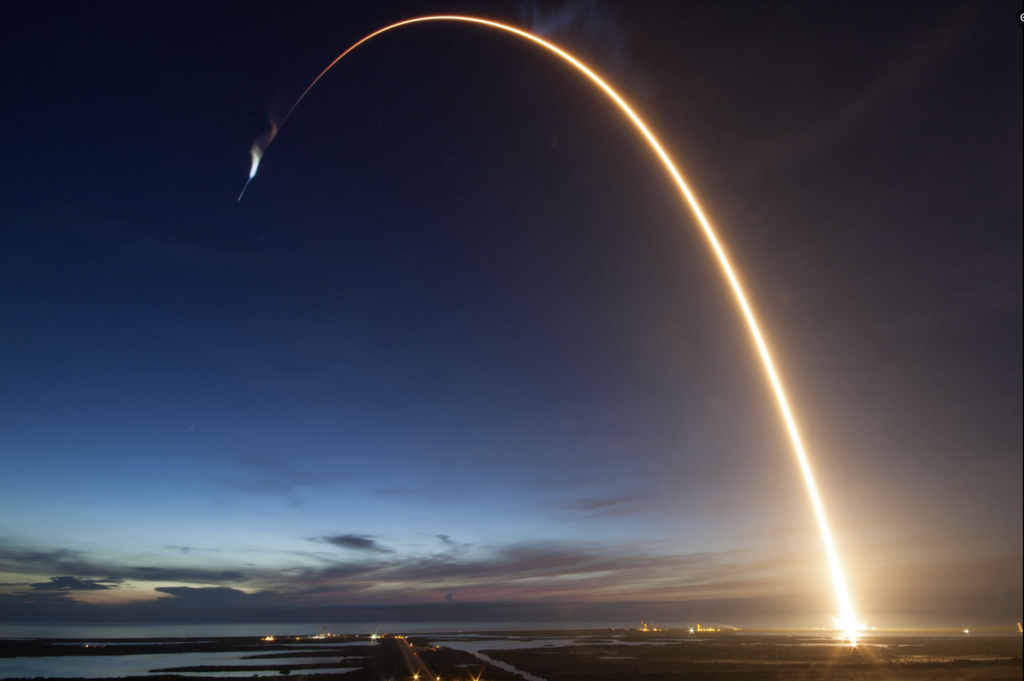 Rocket launch. A bright trail of rocket exhaust creates an arc that rises high in the sky and arcs into a "downward" direction.